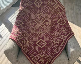 Puede incluir: Una manta de tiro de color burdeos y beige con un patrón geométrico. La manta está colocada sobre un sillón gris.