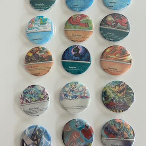 May include: A set of 15 round, colorful buttons featuring Pokemon card art. The buttons are arranged in a grid pattern on a white surface. The buttons feature various Pokemon characters and attacks, including Daring Strike, Bubble, Lunge Out, Sprinkle Water, Energy Assist, Cross-Cut, Unnerve, Stomp Off, Heat Tackle, Prizm Charge,  Deleting Slash, and more.