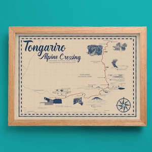 May include: A framed map print of the Tongariro Alpine Crossing in New Zealand. The map has a vintage style with a beige background, blue text, and illustrations. The frame is made of light-coloured wood. The map includes a compass rose.