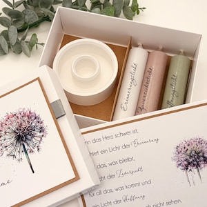 Könnte beinhalten: Eine weiße Geschenkbox mit einem Pusteblumen-Design auf dem Deckel, die einen weißen Kerzenhalter und drei Säulenkerzen in Weiß, Rosa und Grün enthält. Die Kerzen haben Schrifttext. Die Box enthält auch eine Karte mit Text und einer Pusteblumen-Illustration.
