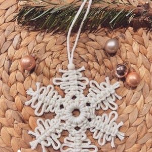 Könnte beinhalten: Ein handgefertigter weißer Makramee-Schneeflocken-Ornament mit einer Schlaufe zum Aufhängen. Die Schneeflocke befindet sich auf einem gewebten, hellbraunen Untergrund, mit kleinen goldenen und roségoldenen Ornamenten und einem Zweig Grün. Der Buchstabe "A" ist unten links.