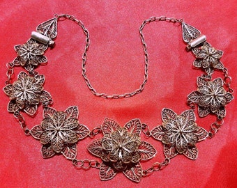 May include: A silver filigree necklace with a delicate chain and floral designs. The necklace is made of intricate silverwork and features a series of connected flower-shaped pendants.