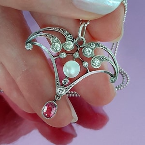 May include: Silver pendant necklace with an Art Nouveau design. The pendant features a central pearl, clear gemstones, and a teardrop-shaped pink gemstone. The necklace is on a delicate silver chain.