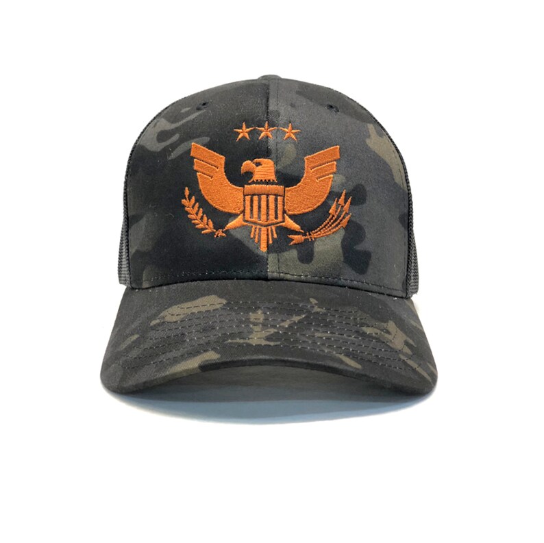 Puede incluir: Gorra de b&eacute;isbol de camuflaje con la parte trasera de malla. La gorra presenta un emblema de &aacute;guila naranja bordado con tres estrellas encima. El sombrero tiene una visera curva y un panel frontal estructurado.