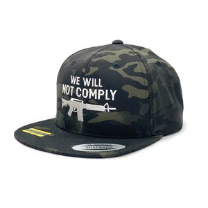May include: A black and green camouflage baseball cap with a white embroidered design that says "We Will Not Comply" and a silhouette of a rifle.