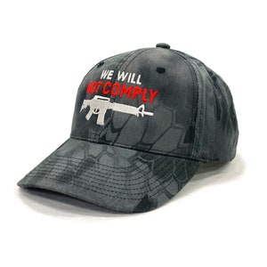 May include: A black and gray camouflage baseball cap with the text "WE WILL NOT COMPLY" in red and white lettering and a white silhouette of a rifle.