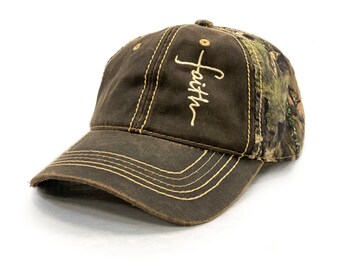 Puede incluir: Una gorra de béisbol marrón con un patrón de camuflaje en la parte posterior. La parte delantera de la gorra tiene la palabra "Faith" bordada en color crema con una cruz sobre la palabra.