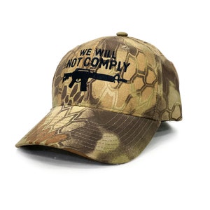 May include: A camouflage baseball cap with the text "WE WILL NOT COMPLY" embroidered in black above a black rifle graphic. The cap features a curved brim and a button on top.