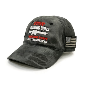 Puede incluir: Una gorra de béisbol de camuflaje negro y gris con el texto "STOP BLAMING GUNS" en rojo y blanco. El texto "TRIGGERS DON'T PULL THEMSELVES" también está en rojo y blanco. Un pequeño parche de la bandera estadounidense está en el lateral de la gorra.