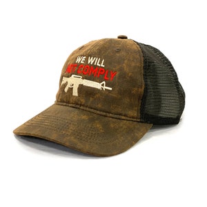 May include: A brown and black trucker hat with a distressed look. The front of the hat features white embroidery of a rifle with the text "WE WILL NOT COMPLY" in red.