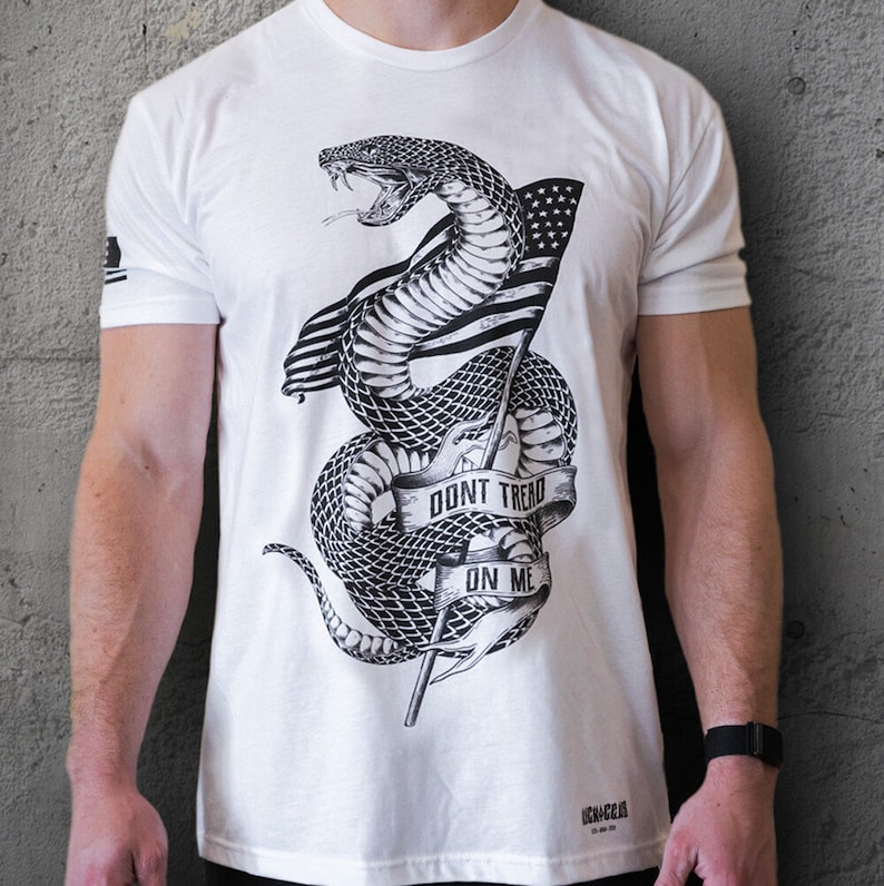 May include: White t-shirt featuring a black and white illustration of a snake wrapped around a flagpole with an American flag. The snake is coiled around the flagpole and the text "Don't Tread On Me" is written on a banner.