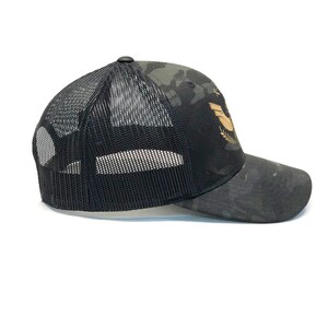 May include: Black and camo trucker hat with a gold embroidered logo of a bird with wings spread and a laurel wreath.