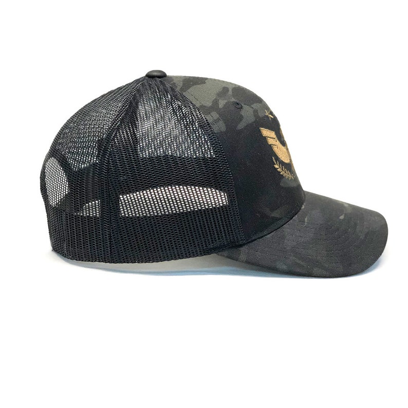 May include: Black and camo trucker hat with a gold embroidered logo of a bird with wings spread and a laurel wreath.