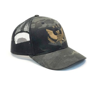 May include: A black and green camouflage trucker hat with a gold embroidered eagle and stars design. The hat has a mesh back.