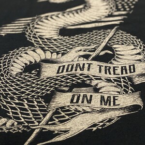 May include: A black t-shirt featuring a graphic of a coiled rattlesnake with the words "Don't Tread On Me" on a banner.