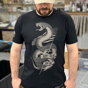 May include: A black t-shirt featuring a graphic of a snake with the words "Don't Tread On Me" on a banner. The snake is coiled around a spear and has stars on its body. The t-shirt is worn with a green and beige baseball cap that says "Join, Or Die."