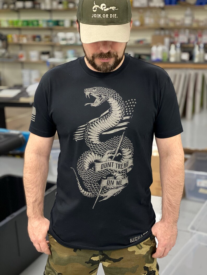 May include: A black t-shirt featuring a graphic of a snake with the words "Don't Tread On Me" on a banner. The snake is coiled around a spear and has stars on its body. The t-shirt is worn with a green and beige baseball cap that says "Join, Or Die."