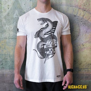 May include: White t-shirt featuring a black and white graphic of a snake wrapped around a flagpole with an American flag. The snake is coiled around the pole and the flag is draped over the snake. The text "Don't Tread On Me" is written on a ribbon around the snake.