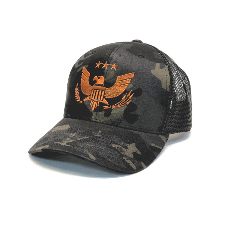 Puede incluir: Gorra de b&eacute;isbol de camuflaje con la parte trasera de malla negra. La parte delantera de la gorra presenta un emblema de &aacute;guila naranja bordado con tres estrellas encima. La gorra tiene una visera curva y una corona estructurada.