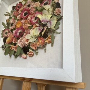 A framed floral display with dried flowers in pink, peach, and cream hues. The flowers are arranged in a circular design against a neutral backdrop, enclosed within a white wooden frame. The artwork is presented on a wooden easel.