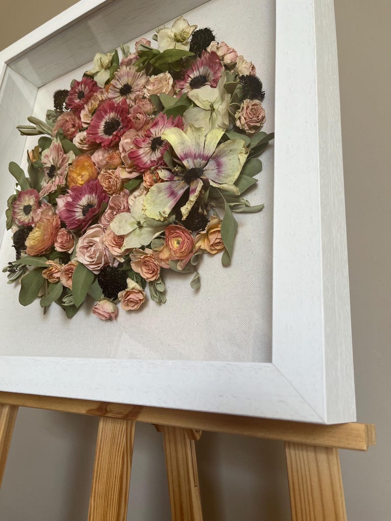 A framed floral display with dried flowers in pink, peach, and cream hues. The flowers are arranged in a circular design against a neutral backdrop, enclosed within a white wooden frame. The artwork is presented on a wooden easel.