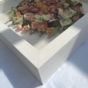 A framed floral arrangement with pink, peach, and green flowers. The flowers are arranged in a circle within a white wooden frame, creating a 3D effect. The square frame is white.