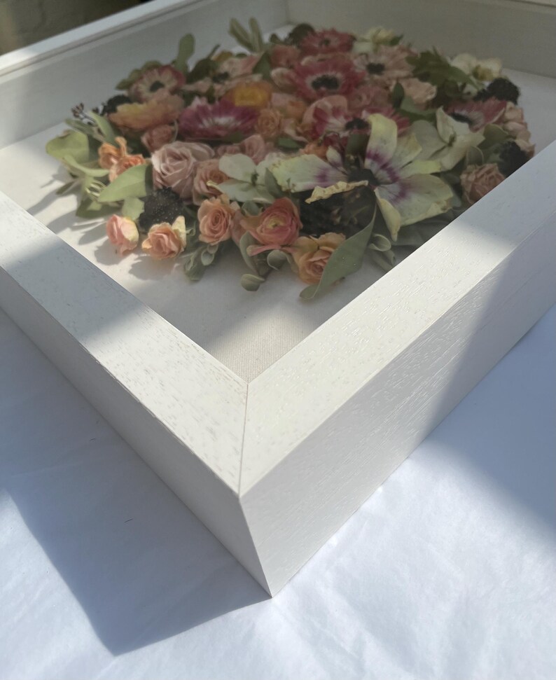 A framed floral arrangement with pink, peach, and green flowers. The flowers are arranged in a circle within a white wooden frame, creating a 3D effect. The square frame is white.