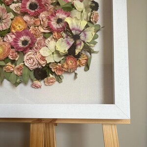 A framed floral arrangement of dried flowers in pink, peach, and yellow hues. The flowers are arranged in a circular, ‘bloom cluster’ design against a neutral backdrop, enclosed within a white wooden frame. The artwork is displayed on a wooden easel.