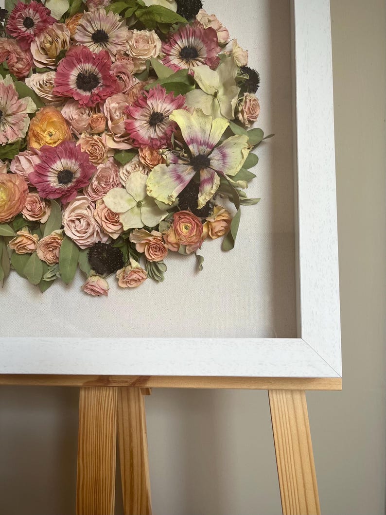 A framed floral arrangement of dried flowers in pink, peach, and yellow hues. The flowers are arranged in a circular, ‘bloom cluster’ design against a neutral backdrop, enclosed within a white wooden frame. The artwork is displayed on a wooden easel.