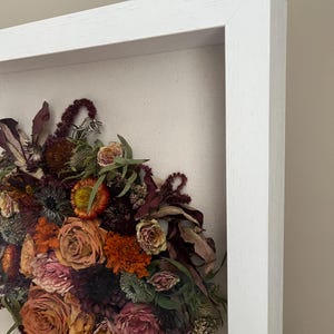 A framed floral arrangement of dried flowers in shades of orange, pink, and burgundy. The flowers are set against a neutral canvas background and enclosed in a white wooden frame. The arrangement includes roses and other dried botanicals.