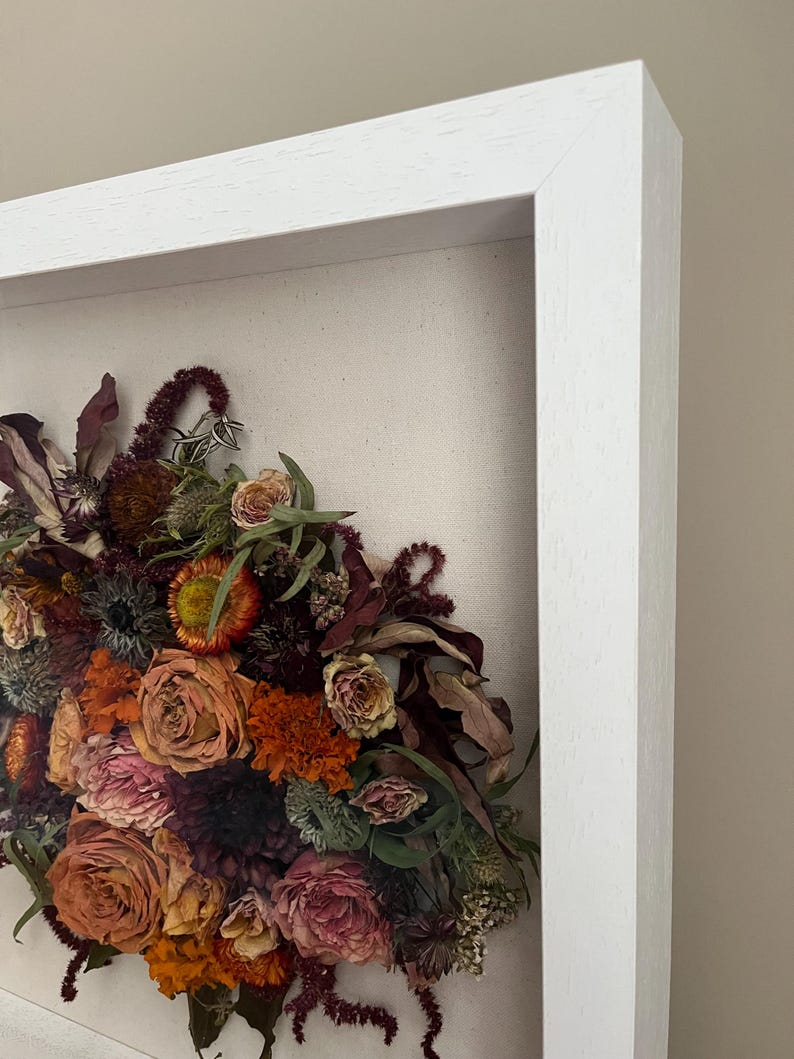A framed floral arrangement of dried flowers in shades of orange, pink, and burgundy. The flowers are set against a neutral canvas background and enclosed in a white wooden frame. The arrangement includes roses and other dried botanicals.