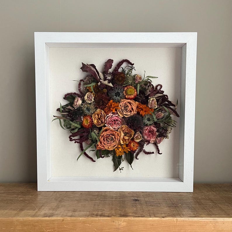 A framed display of dried flowers in a white square frame. The arrangement features a variety of blooms in shades of orange, pink, and burgundy, set against a cream-coloured background. The flowers are arranged in a circular shape.