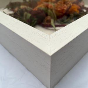 Close-up of a white wooden shadow box frame with a glass front. The frame contains dried flowers in shades of orange, red, and green. The corner of the frame is visible, showing the wood grain texture.