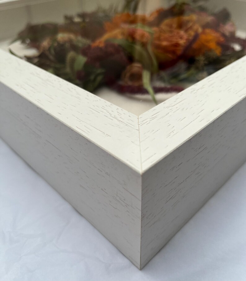 Close-up of a white wooden shadow box frame with a glass front. The frame contains dried flowers in shades of orange, red, and green. The corner of the frame is visible, showing the wood grain texture.