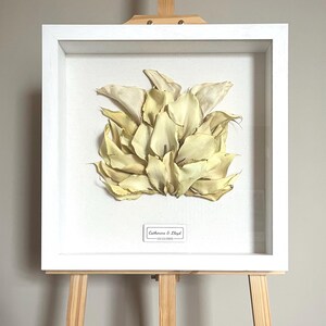 May include: A framed floral art piece featuring a cluster of dried, cream-colored petals. The artwork is encased in a white, square shadow box frame and displayed on a wooden easel. A small label at the bottom reads "Catherine & Lloyd - 13/12/2015."