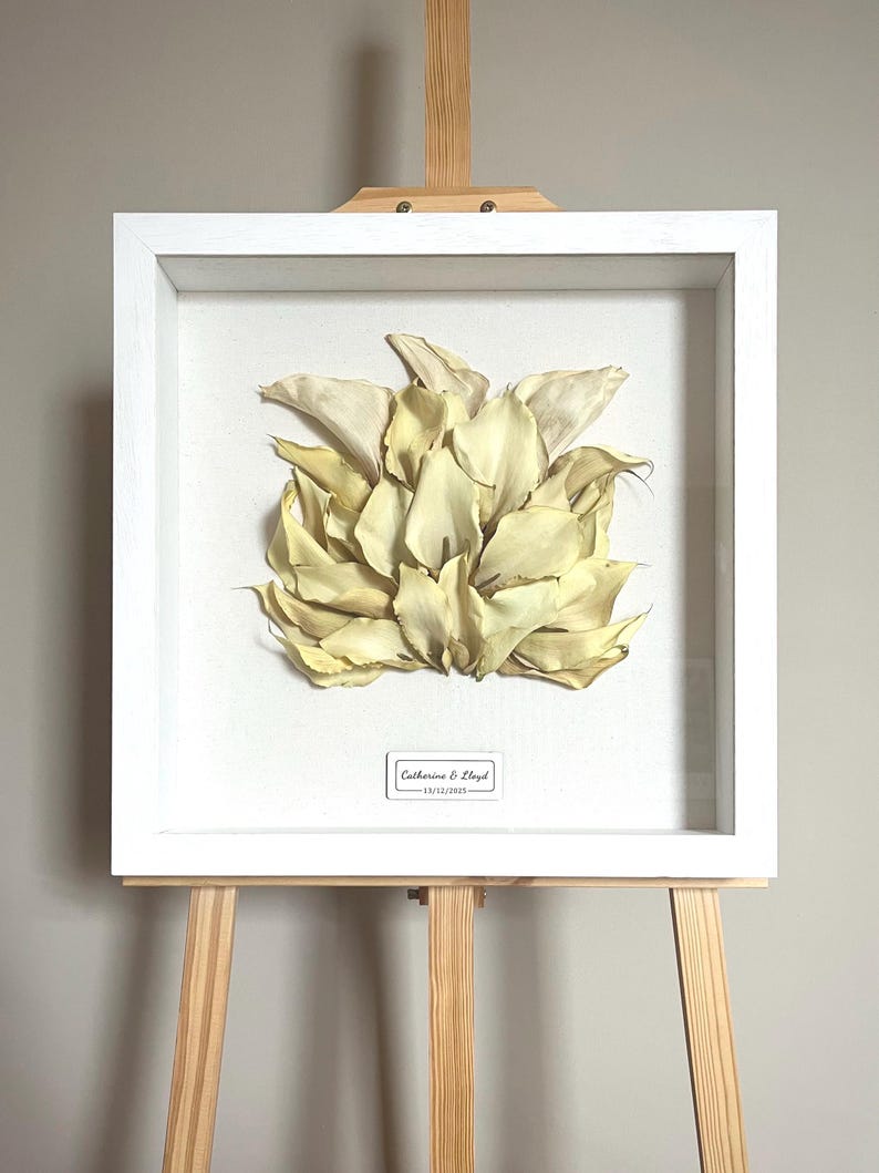 May include: A framed floral art piece featuring a cluster of dried, cream-colored petals. The artwork is encased in a white, square shadow box frame and displayed on a wooden easel. A small label at the bottom reads "Catherine & Lloyd - 13/12/2015."