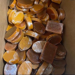 May include: A collection of numerous oval and rectangular bars of soap, each individually wrapped in clear plastic film. The bars vary in shades of amber, golden yellow, and deep brown, creating a dense, layered arrangement within a container.