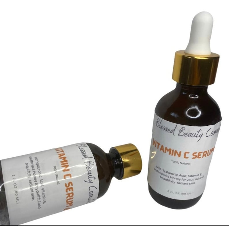 K&ouml;nnte beinhalten: Zwei braune Glasflaschen mit goldenen Kappen und Etiketten mit der Aufschrift "Blessed Beauty Cosmetics Vitamin C Serum 100% Nat&uuml;rlich" mit dem Text "mit Hyalurons&auml;ure, Vitamin E und Manuka-Honig f&uuml;r jugendliche und strahlend sch&ouml;ne Haut." Die Flaschen sind 2 fl oz (60 ml).