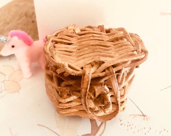 May include: A small, woven wicker basket with a lid. The basket is light brown and has a woven pattern. The basket is sitting on a white surface with a pink unicorn toy in the background.