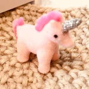 May include: A small pink plush unicorn toy with a silver glitter horn. The unicorn is sitting on a brown crocheted surface.
