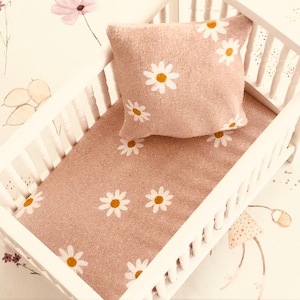 May include: A white cot with a pink and white daisy patterned bedding set. The cot has a white wooden frame and a pink and white daisy patterned mattress. The bedding set includes a pillow with a pink and white daisy patterned cover.