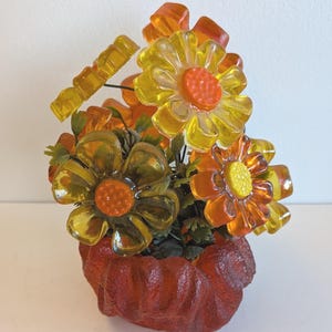 May include: A decorative arrangement of translucent yellow and orange acrylic flowers with textured centres. The flowers are attached to thin green wire stems with small green leaves, all set within a textured, dark orange, bowl-shaped base.