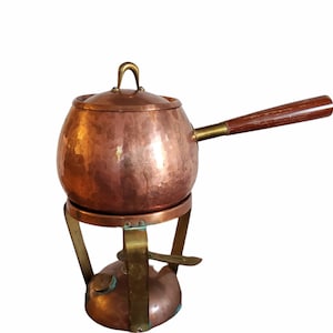 May include: A copper fondue pot with a hammered finish and a wooden handle. The pot sits on a brass stand with a burner.