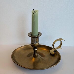 Peut inclure: Un bougeoir en laiton vintage avec une base ronde et une poignée. Une bougie verte est placée au centre. Le bougeoir a un aspect terni. La bougie n'est pas allumée.