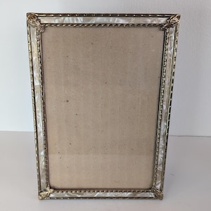 May include: A rectangular picture frame featuring a metallic gold-toned border with intricate rope-like detailing and decorative corner accents. The frame includes a translucent, iridescent white inlay along the inner edge and a plain tan backing.