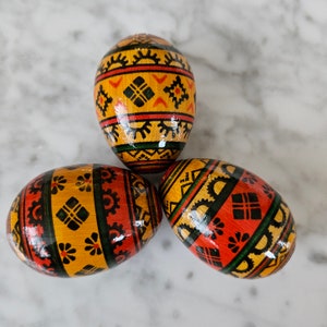 Peut inclure: Trois œufs de Pâques peints à la main avec des motifs géométriques dans des tons de rouge, jaune, noir et vert.