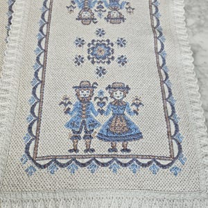 May include: Cream-coloured table runner with a detailed woven design. The runner features a repeating pattern of figures in blue and brown, with floral and geometric accents. The edges are finished with a scalloped border and fringe.