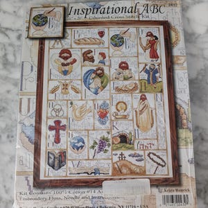 May include: A packaged cross-stitch kit titled "Inspirational ABC." The kit features a framed design with religious imagery and text. The design measures 16 x 20 inches and includes embroidery floss and a needle.