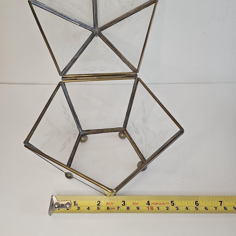 May include: A clear glass geometric box with a brass frame and etched details. The box is open and has a hinged lid. The box is approximately 25 cm wide.