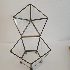 May include: A clear glass geometric box with a lid. The box has a hexagonal base and a pyramid-shaped lid. The box is made of clear glass with a frosted finish. The box is sitting on a white surface.